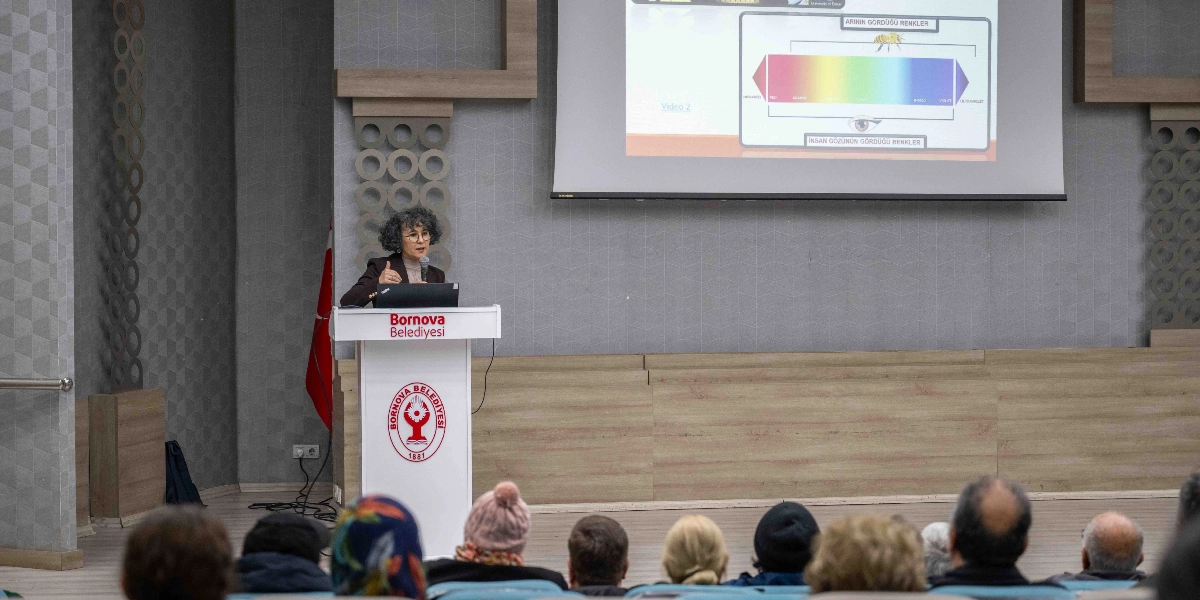 Kıvırcık saçlı, gözlüklü bir kadın, üzerinde Bornova Belediyesi logosu bulunan bir kürsüde konuşma yapıyor. Kadının arkasındaki projeksiyon perdesinde, arıların ve insanların renk algısını karşılaştıran şematik bir görsel yer alıyor. İzleyiciler, salondaki koltuklarda oturarak sunumu takip ediyor.