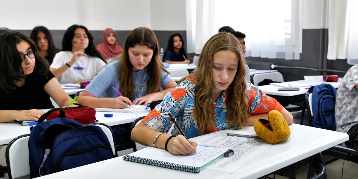 Sınıf ortamında sıralarında oturan ve ders çalışan öğrenciler. Ön planda, üzerinde mavi ve turuncu yaprak desenli bir gömlek olan, uzun sarı saçlı bir kız öğrenci not alıyor. Yanında ve arkasında farklı kıyafetler giymiş, dikkatle dersi takip eden veya yazı yazan diğer öğrenciler bulunuyor. Masaların üzerinde defterler, kalemler ve kalemlikler yer alıyor.