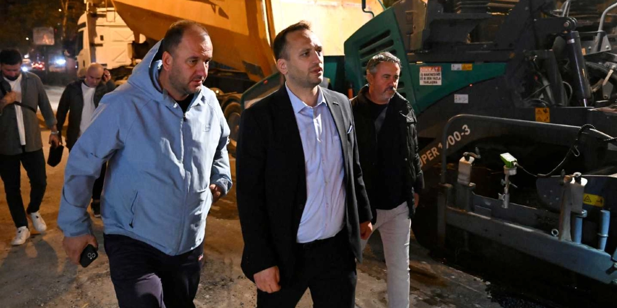 Gece saatlerinde Bornova'da devam eden yol çalışması alanında, açık mavi kapüşonlu bir ceket giyen adam, yanında lacivert takım elbiseli bir yönetici ve arkasında siyah montlu bir başka kişiyle birlikte yürürken görülüyor. Arka planda büyük bir asfalt serme makinesi ve sarı renkli bir kamyon bulunuyor.