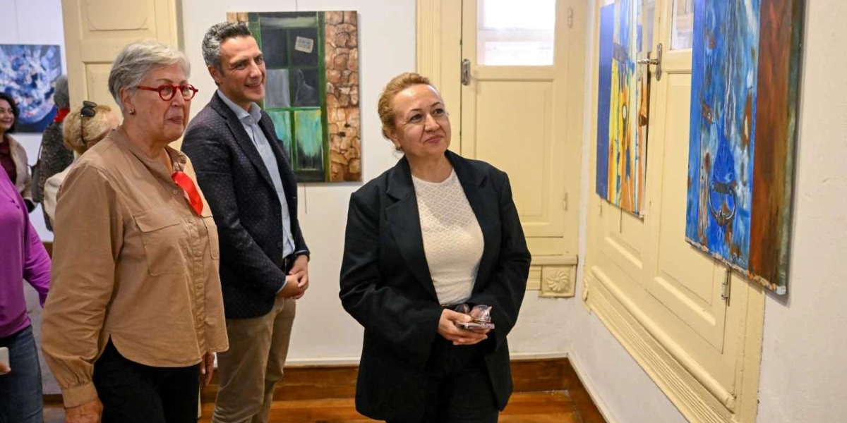 Bornova'daki bir sanat galerisinde, duvarda asılı olan mavi tonlarındaki soyut bir tabloyu inceleyen üç kişi. En önde, siyah ceketli ve beyaz bluzlu bir kadın tabloya bakarken, hemen arkasında lacivert ceketli bir erkek ve kırmızı gözlüklü, bej gömlekli bir kadın ona eşlik ediyor. Arka planda ise başka tablolar ve ahşap pencereli bir duvar detayı görülüyor.