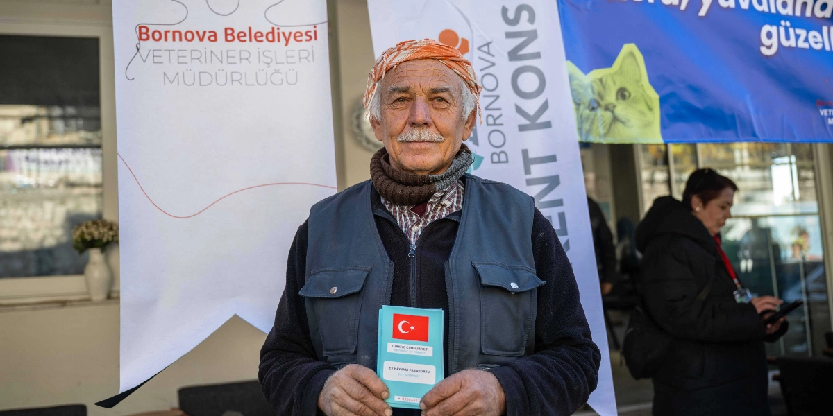 Başına turuncu bir yazma bağlamış, gri bıyıklı, koyu renk yelek giyen yaşlı bir adam, elinde Türkiye Cumhuriyeti Ev Hayvanı Pasaportu tutarak kameraya bakıyor. Arka planda Bornova Belediyesi Veteriner İşleri Müdürlüğü ve Bornova Kent Konseyi logolarının bulunduğu afişler yer alıyor.
