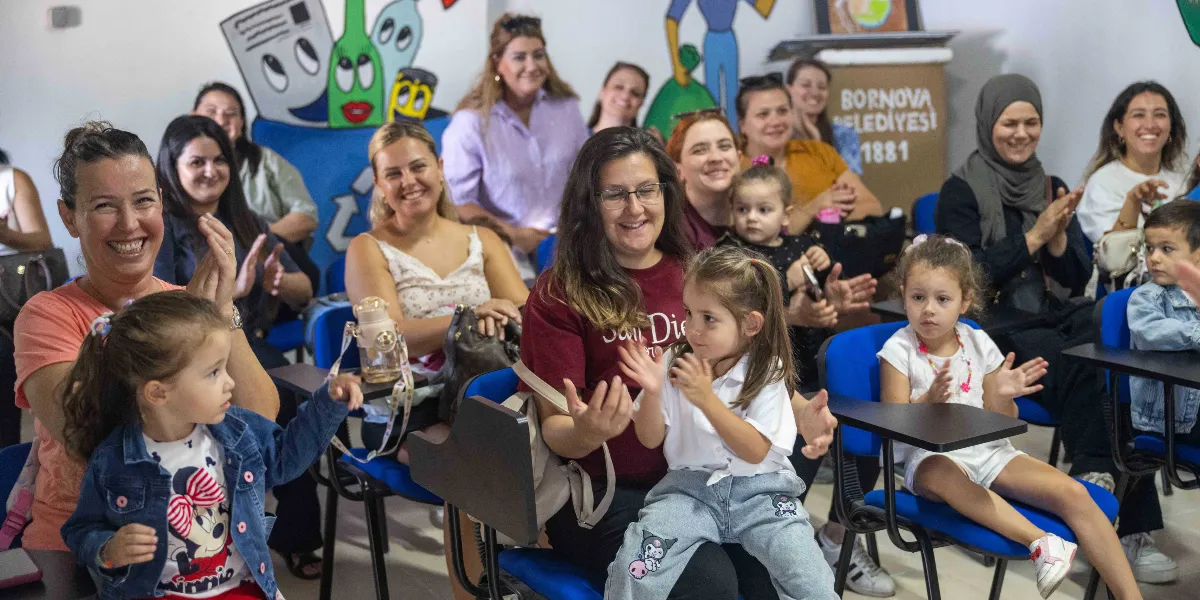 İç mekan bir etkinlik alanında, aralarında çocukların da bulunduğu bir grup kadın neşeyle alkışlıyor. Katılımcılar mavi plastik sandalyelerde oturuyor, ön planda kucaklarında çocukları olan anneler dikkat çekiyor. Arka planda, üzerinde 