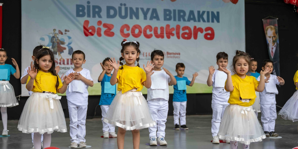 23 Nisan Ulusal Egemenlik ve Çocuk Bayramı kutlamaları kapsamında sahnede dans eden bir grup çocuk. Çocukların bir kısmı sarı tişört ve beyaz tül etek, bir kısmı ise beyaz veya mavi tişört ve beyaz pantolon giymiş durumda. Arka planda üzerinde 