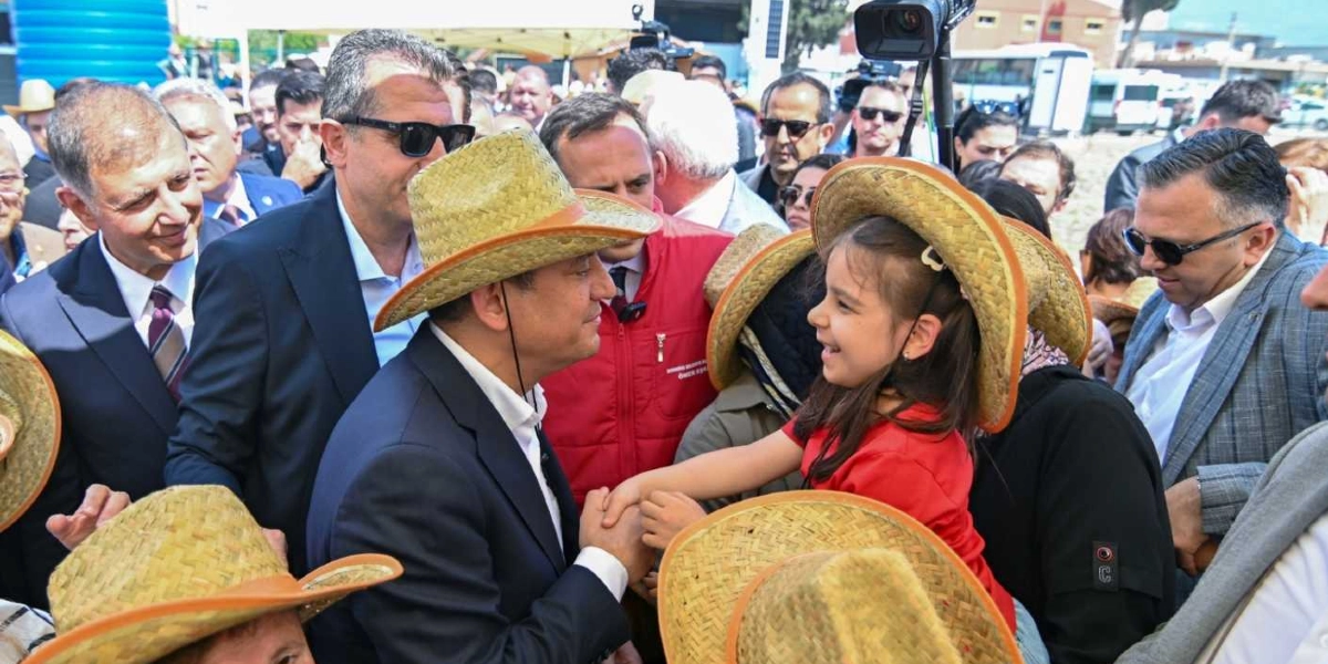 CHP Genel Başkanı Özgür Özel, başında hasır bir şapka ile kalabalık bir ortamda, kırmızı tişörtlü ve yine hasır şapkalı küçük bir kız çocuğuyla el sıkışarak gülümsüyor. Çevresinde takım elbiseli ve güneş gözlüklü birçok kişinin yer aldığı dış mekan etkinliğinde, katılımcıların birçoğu da benzer hasır şapkalar takıyor.