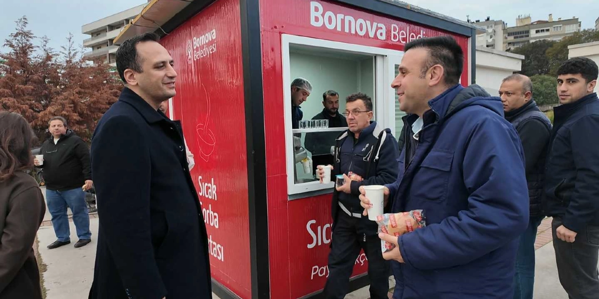 Bornova Belediyesi'ne ait kırmızı renkli bir çorba dağıtım standının önünde, ellerinde karton bardaklarla çorba içen ve sohbet eden bir grup insan yer alıyor. Standın üzerinde 
