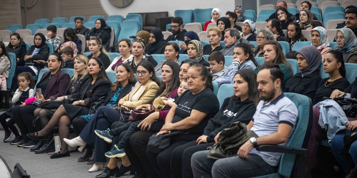 Konferans salonunda yan yana dizilmiş mavi koltuklarda oturan, büyük çoğunluğu kadınlardan oluşan kalabalık bir izleyici grubu. Katılımcılar dikkatle karşıya bakarak bir etkinliği takip ediyor; aralarında çocukların da bulunduğu izleyicilerin bir kısmı başörtülü, bir kısmı ise günlük kıyafetler içinde yer alıyor. Ön sıralarda oturan yetişkinler ve kucaklarında veya yanlarında çocukları olan ebeveynler, ciddiyetle sahneye odaklanmış durumda.