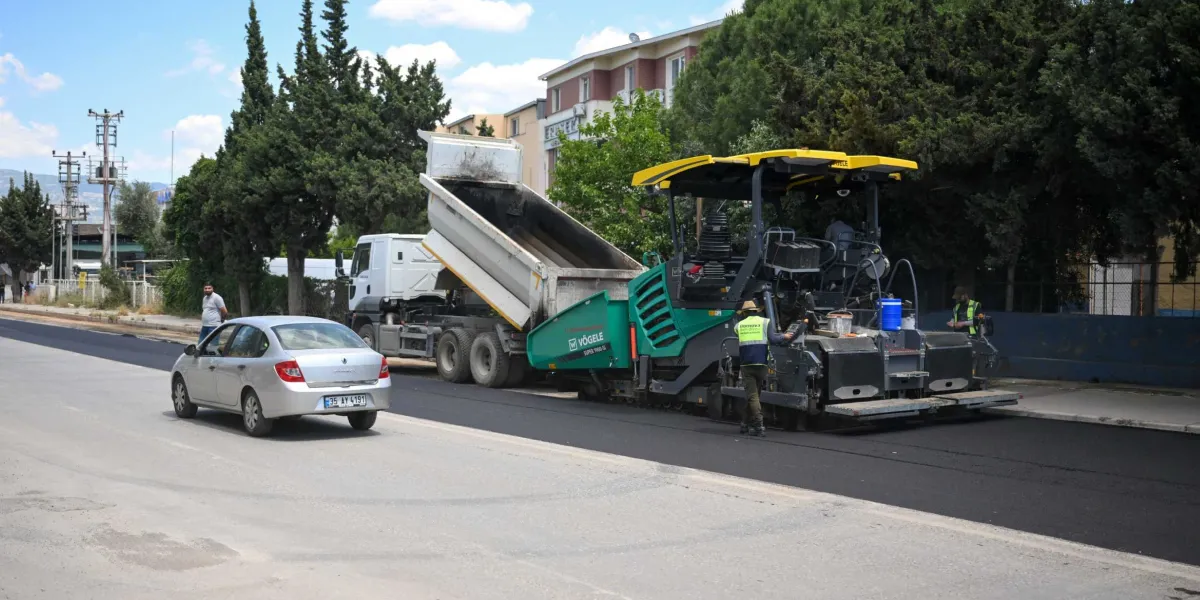 Pınar Caddesi'nde devam eden yol yenileme çalışmaları kapsamında, damperli bir kamyonun asfalt serici makineye malzeme boşalttığı görülüyor. Yeşil ve siyah renkli Vögele marka asfalt serici makinenin başında sarı yelekli bir operatör bulunurken, çalışma alanının hemen yanında gümüş renkli bir binek otomobil ilerliyor. Arka planda ağaçlar ve yerleşim yerleri yer alıyor.