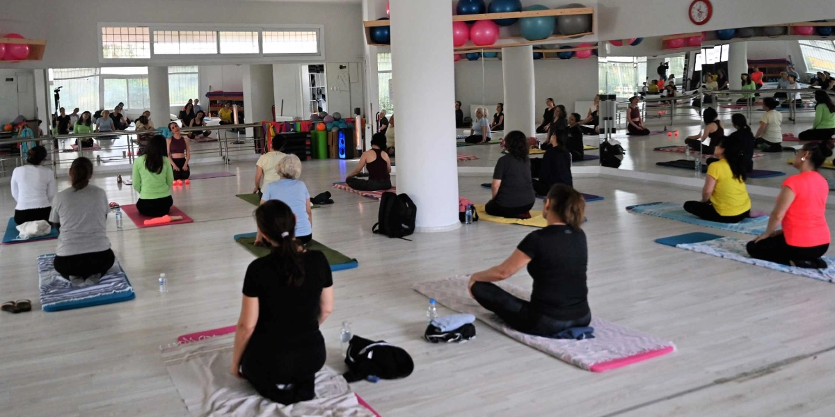 Aydınlık ve geniş bir spor salonunda, yoga matları üzerinde oturan kadınlar eğitmen eşliğinde yoga yapıyor. Katılımcılar farklı renklerde spor kıyafetleri giymiş, sırtları kameraya dönük şekilde yere oturmuş veya bağdaş kurmuş durumda. Salonun bir duvarı tamamen aynalarla kaplı olduğu için içerideki kalabalık ve ortam yansımalarla daha geniş görünüyor. Tavan kısmındaki raflarda renkli pilates topları dizili ve salonun genelinde huzurlu bir çalışma ortamı hakim.