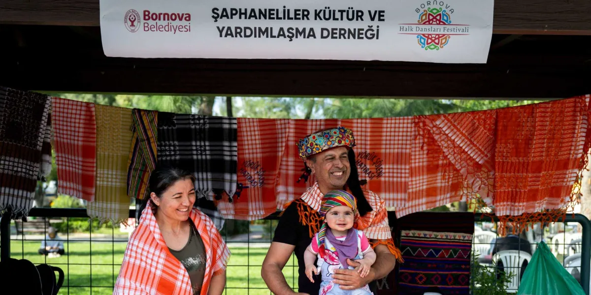 Bornova Belediyesi Şaphaneliler Kültür ve Yardımlaşma Derneği standının önünde, omuzlarında turuncu-beyaz desenli yöresel şallar taşıyan bir kadın ve bir erkek ile erkeğin kucağında renkli bir başlık takan bir bebek gülümsüyor. Arka planda ipe asılmış çeşitli renk ve desenlerde şallar sergileniyor.