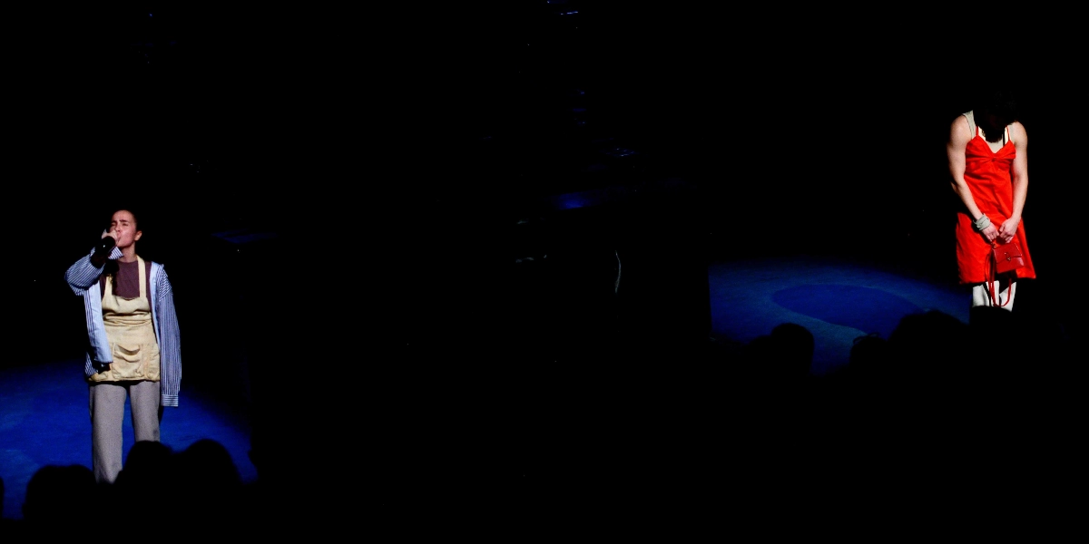 Karanlık bir sahnede iki oyuncu spot ışığı altında duruyor. Soldaki oyuncu çizgili bir gömlek ve önlük giymiş, elindeki şişeden içmektedir. Sağdaki oyuncu ise kırmızı askılı bir elbise giymiş, başı öne eğik şekilde elindeki kırmızı çantaya bakmaktadır.