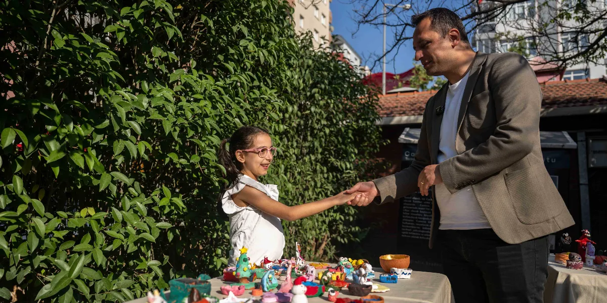 Açık havada, yeşil yapraklı bir ağacın önünde kurulan stantta, gözlüklü küçük bir kız çocuğu ile gri ceketli bir adam el sıkışıyor. Masanın üzerinde çocukların yaptığı renkli ve çeşitli kil figürler sergileniyor.