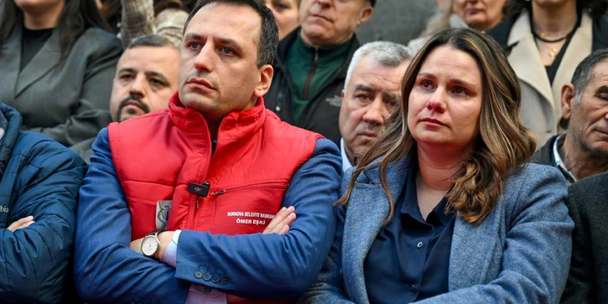 Bornova Belediye Başkanı Ömer Eşki, üzerinde kırmızı yeleğiyle kollarını kavuşturmuş bir şekilde otururken, yanında duygusal bir ifadeyle ileriye bakan bir kadın ve çevresinde dikkatle dinleyen bir kalabalık yer alıyor.