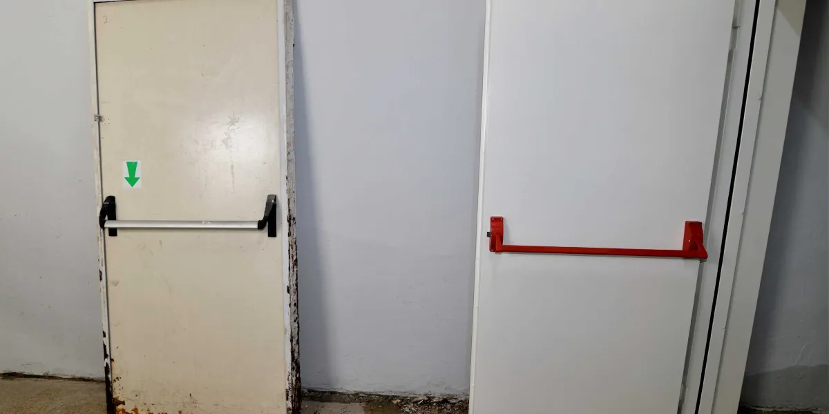 Gri bir duvarın önünde yan yana duran iki adet beyaz acil çıkış kapısı. Soldaki kapının üzerinde aşağıyı gösteren yeşil bir ok işareti ve gümüş renkli bir panik bar bulunurken, sağdaki kapıda kırmızı renkli bir panik bar yer alıyor.