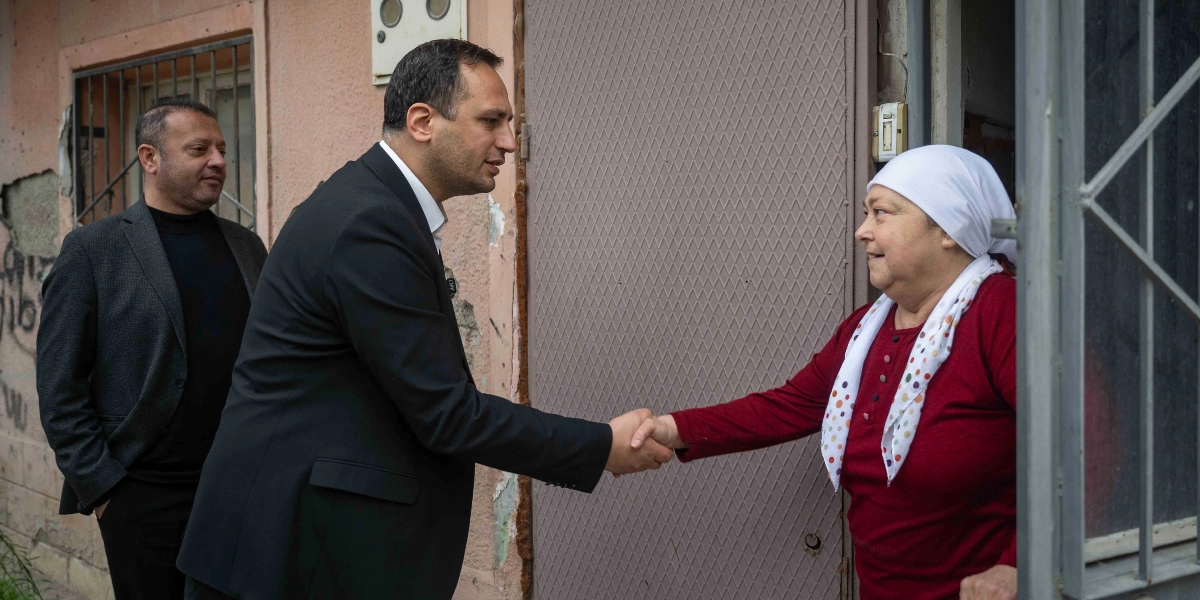 Koyu renk takım elbiseli bir erkek, evinin kapısında duran beyaz başörtülü ve kırmızı bluzlu bir kadınla tokalaşıyor. Arkalarında ise gri ceketli başka bir erkek daha bulunuyor.