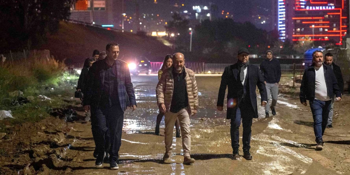 Gece vakti, çamurlu ve engebeli bir arazide yürüyen bir grup insan. Ön planda, koyu renkli ceketli, bej şişme montlu ve siyah kabanlı erkeklerden oluşan bir heyet, yan yana yürüyerek incelemelerde bulunuyor. Arka planda ise şehrin ışıkları ve üzerinde kırmızı ışıklandırmalı 