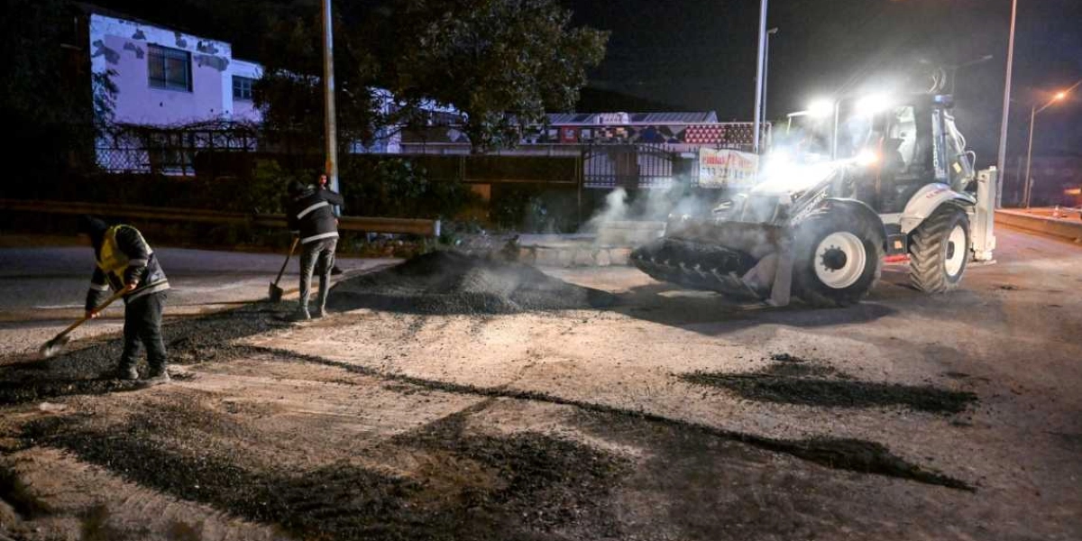 Gece vakti Bornova'da bir yol çalışması sırasında, iş makineleriyle dökülen sıcak asfaltı küreklerle düzelten iki işçi ve arkalarında bekleyen bir kepçe görülüyor.