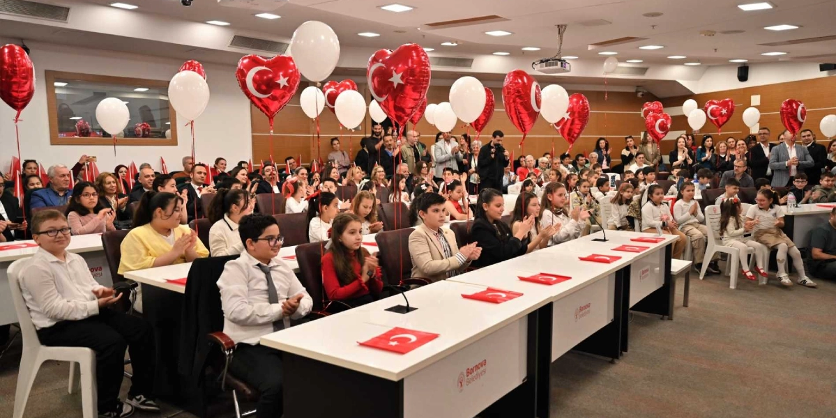 Bornova Belediyesi'nin düzenlediği bir etkinlikte, sıralarda oturan çocuklar ve arkalarında onları izleyen yetişkinler alkışlıyor. Salon, üzerinde Türk bayrağı baskılı kalp şeklinde kırmızı balonlar ve beyaz balonlarla süslenmiş durumda. Masaların üzerinde küçük Türk bayrakları yer alıyor ve ön planda şık giyimli çocuklar dikkat çekiyor.