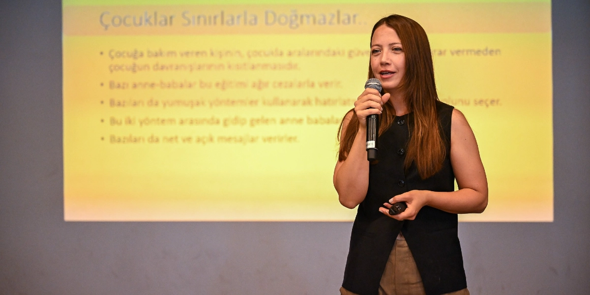 Uzun kahverengi saçlı bir kadın, elinde mikrofonla bir sunum yapıyor. Arkasındaki yansıtıcı perdede 