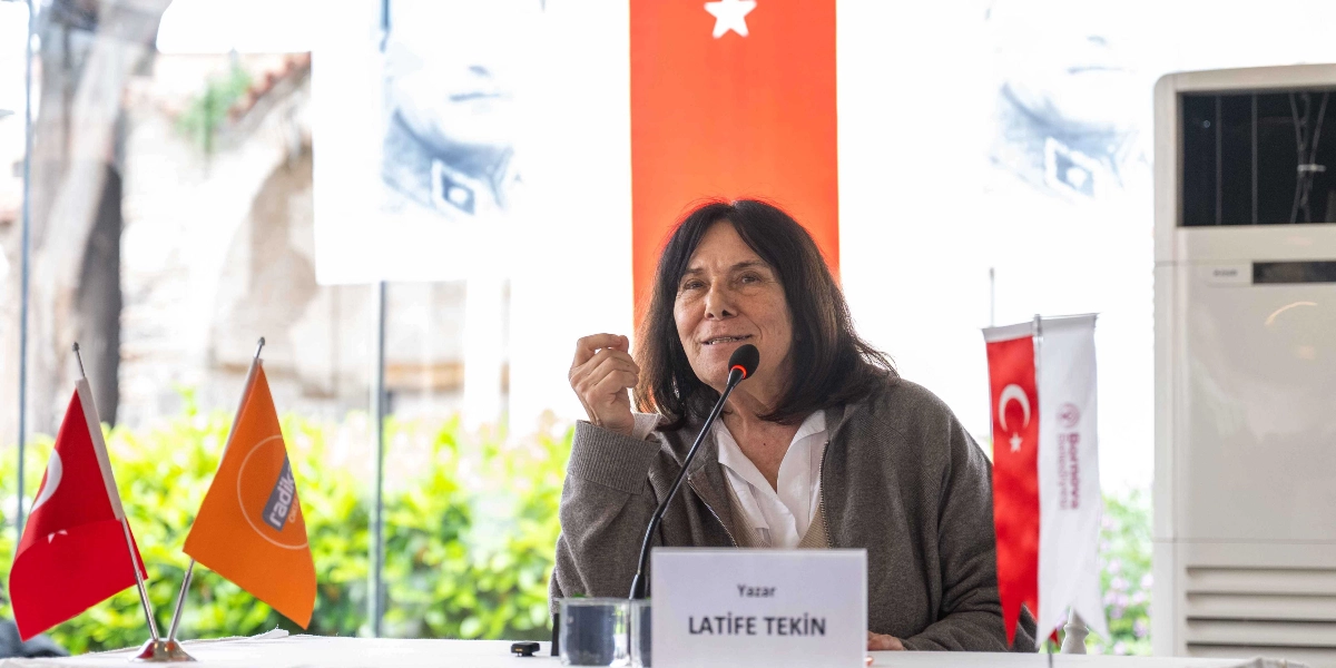 Yazar Latife Tekin, üzerinde isminin ve unvanının yazılı olduğu bir isimliğin bulunduğu masada, mikrofon karşısında konuşma yapıyor. Arka planda Türk bayrağı ve üzerinde Mustafa Kemal Atatürk'ün silüetinin bulunduğu bir flama yer alıyor. Masanın üzerinde ayrıca küçük bir Türk bayrağı, turuncu renkli Radikal Gazetesi flaması ve Bornova Belediyesi logolu bir bayrak bulunuyor.