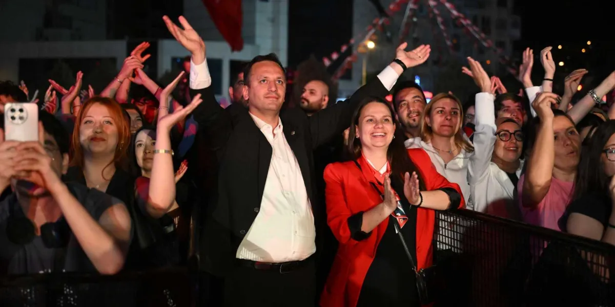 Gece vakti açık havada düzenlenen bir etkinlikte, ellerini havaya kaldırarak alkışlayan ve coşkuyla gülümseyen kalabalık bir grup insan. Ön planda siyah ceketli ve beyaz gömlekli bir erkek ile kırmızı ceketli bir kadın yer alıyor. İzleyicilerin bir kısmı telefonlarıyla çekim yaparken, arka planda ise etkinlik alanını çevreleyen metal bariyerler ve bulanık ışıklar görülüyor.