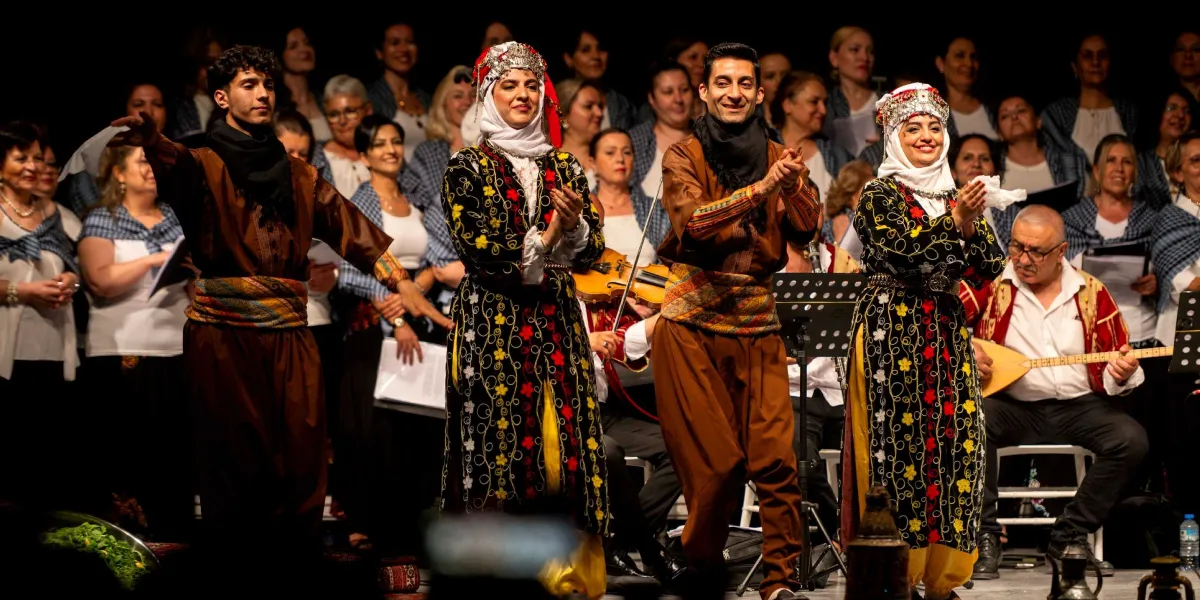 Sahne üzerinde geleneksel kıyafetler giymiş iki erkek ve iki kadın dansçı, halay çekerken elleriyle tempo tutuyor. Erkekler kahverengi şalvar ve kuşaklı üstler giymiş, kadınlar ise siyah üzerine renkli çiçek desenli uzun elbiseler ve beyaz başlıklar takmış durumda. Dansçıların arkasında, beyaz tişört ve omuzlarında mavi desenli şallar bulunan geniş bir koro grubu yer alıyor. Sağ tarafta ise saz çalan bir müzisyen ve keman çalan bir başka kişi görülüyor. Sahne ışıkları altında gerçekleşen bu kültürel etkinlikte, katılımcılar neşeli bir şekilde performanslarını sergiliyor.