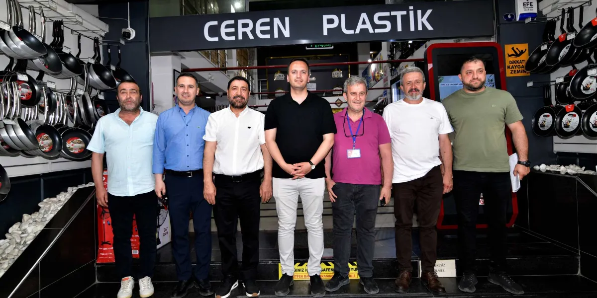 Ceren Plastik tabelasının altında, bir mağazanın girişinde yan yana duran yedi erkek. Arka planda duvarlarda asılı tava ve tencere çeşitleri görülüyor. Başkan Eşki ve beraberindeki heyet, mağaza ziyareti sırasında objektiflere poz veriyor.