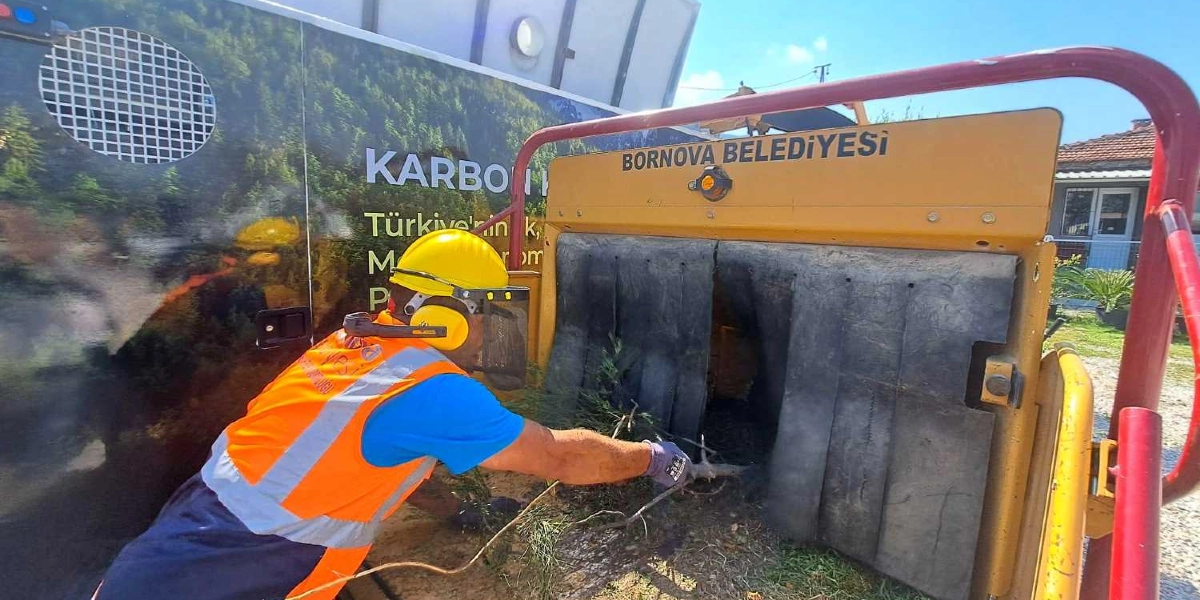 Bornova Belediyesi logolu bir dal öğütme makinesinin başında, sarı baret, koruyucu yüz siperi, kulaklık ve turuncu reflektörlü iş yeleği giymiş bir belediye çalışanı, topladığı ağaç dallarını makinenin girişine yerleştiriyor. Makinenin üzerinde 