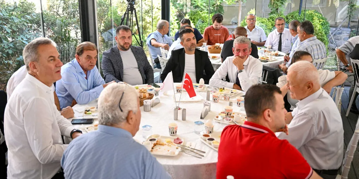 Dramalılar Köşkü'nde düzenlenen bir toplantıda, yuvarlak bir masa etrafında toplanmış, çoğunluğu erkeklerden oluşan bir grup katılımcı kahvaltı yaparken görülüyor. Masanın ortasında Türk bayrağı ve Bornova Belediyesi'ni temsil eden bir flama yer alıyor. Katılımcılar günlük ve resmi kıyafetlerle oturmuş, birbirleriyle sohbet ediyorlar. Arka planda cam duvarlarla çevrili aydınlık bir mekan ve dışarıdaki yeşil bitkiler seçiliyor.