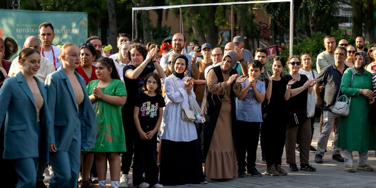 Açık havada düzenlenen bir etkinlikte, mavi ceketli iki dansçıyı izleyen ve alkışlayan farklı yaş gruplarından oluşan kalabalık bir grup. İzleyiciler arasında yeşil elbiseli bir kız çocuğu, beyaz bluzlu ve siyah etekli bir kadın, başörtülü bir kadın ve çeşitli kıyafetler giymiş yetişkinler yer alıyor. Arka planda yeşil bir etkinlik panosu ve ağaçlar görülüyor.