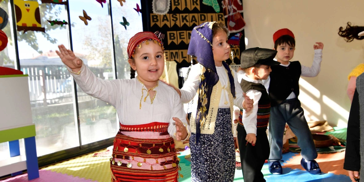 Yerli Malı Haftası kutlamaları kapsamında geleneksel kıyafetler giymiş dört çocuk, bir sınıf ortamında dans ediyor. Ön planda, başında altın işlemeli bir başlık ve belinde yine altın süslemeli kırmızı bir kuşak olan kız çocuğu gülümsüyor. Yanında lacivert yazmalı ve işlemeli yelekli bir kız çocuğu, arkalarında ise siyah yelekli ve fesli iki erkek çocuk yer alıyor. Arka planda duvarda üzerinde 