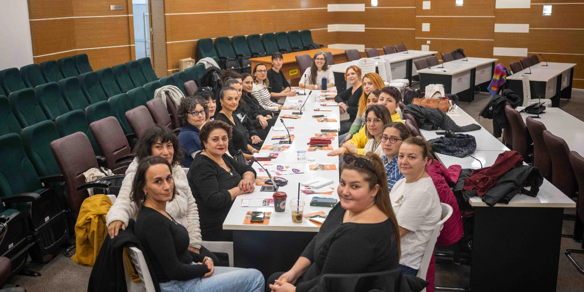 Modern bir toplantı salonunda, uzun bir masanın etrafında oturmuş, çoğunluğu kadınlardan oluşan bir grup katılımcı kameraya gülümsüyor. Masanın üzerinde broşürler, not defterleri, mikrofonlar ve içecekler bulunuyor. Arka planda ise kademeli olarak yükselen yeşil renkli konferans koltukları yer alıyor.