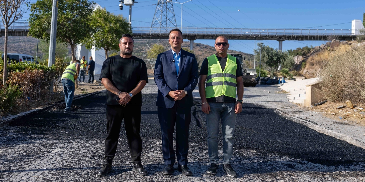 Açık havada, yeni asfalt dökülmüş bir yol üzerinde yan yana duran üç erkek. Ortadaki kişi lacivert takım elbiseli, sağındaki ve solundaki kişiler ise günlük kıyafetler giymiş durumda; sağdaki kişi ayrıca neon sarı bir iş yeleği takıyor. Arka planda yol çalışması yapan diğer belediye işçileri, ağaçlar, bir yaya üst geçidi ve elektrik direkleri görülüyor.