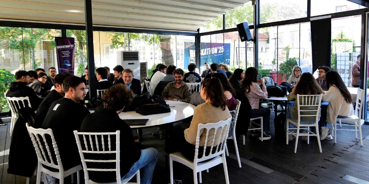 Camla çevrili aydınlık bir salonda, beyaz yuvarlak masaların etrafında toplanmış gençlerin katıldığı bir hackathon etkinliği. Katılımcılar dizüstü bilgisayarlarıyla çalışırken, arka planda üzerinde 