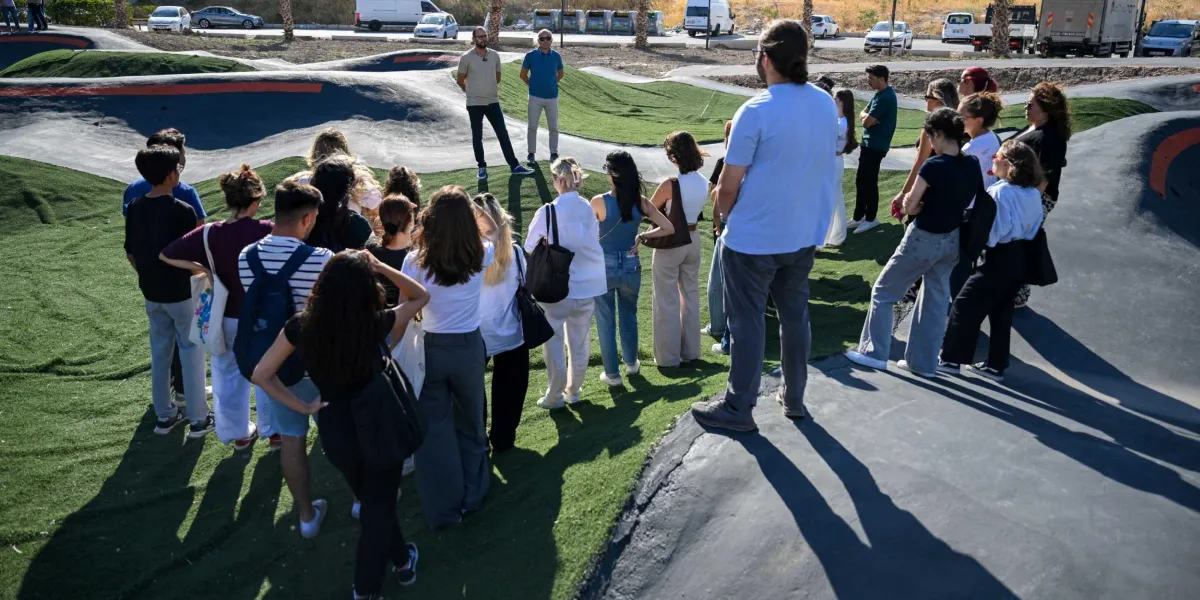 Bornova'daki bir pump track parkurunda toplanmış, parkuru inceleyen kalabalık bir grup insan. Parkurun başında duran iki erkek yetkili, çevresindeki katılımcılara bilgi veriyor. Parkur, asfalt zeminli engebeli yollardan ve aralarda yer alan yapay yeşil çim alanlardan oluşuyor. Arka planda ise parkın yanındaki yoldan geçen araçlar görülüyor.