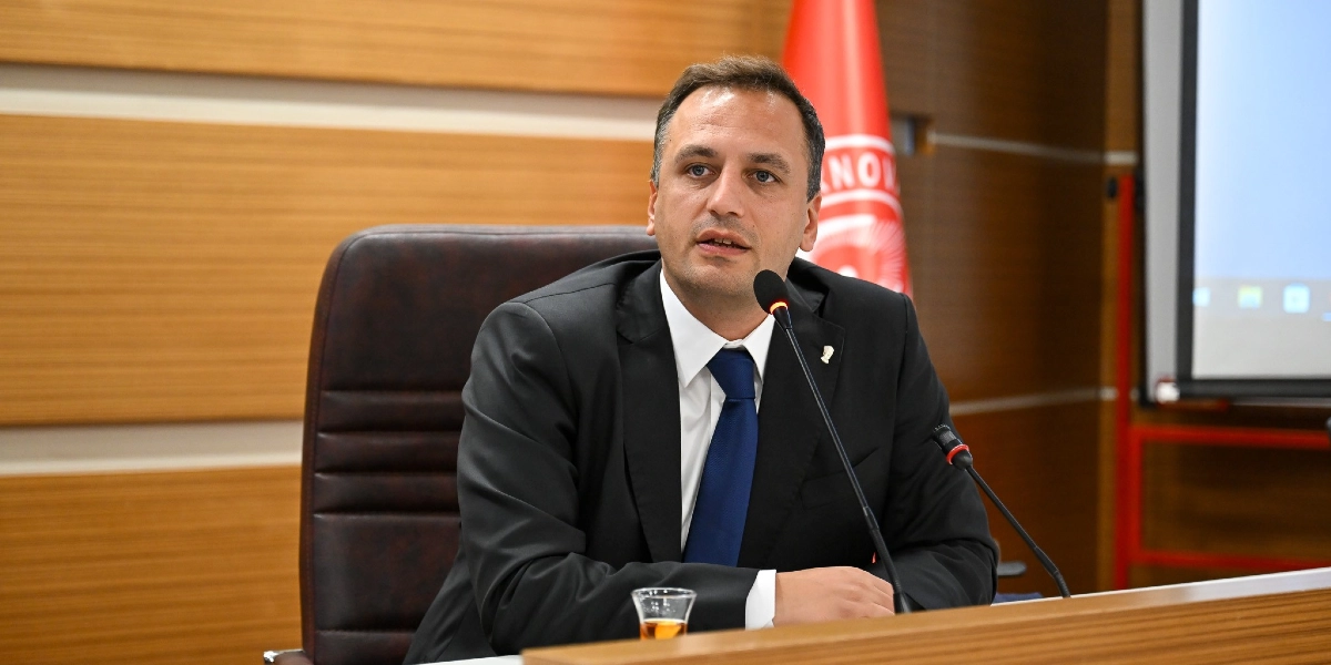 Siyah takım elbiseli, beyaz gömlekli ve lacivert kravatlı bir erkek, ahşap panelli bir toplantı salonunda masada oturarak konuşma yapıyor. Önünde bir mikrofon ve ince belli bir bardakta çay bulunuyor. Arka planda Bornova Belediyesi'ne ait olduğu anlaşılan kırmızı renkli bir bayrak yer alıyor.
