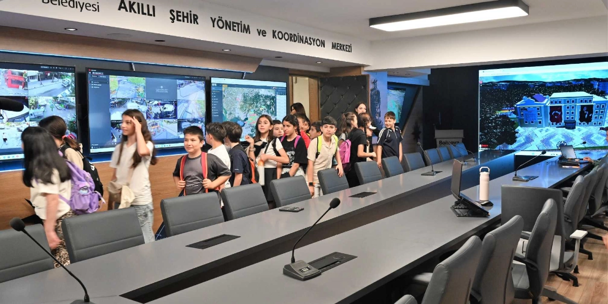 Bornova Belediyesi Akıllı Şehir Yönetim ve Koordinasyon Merkezi'ni ziyaret eden bir grup ilköğretim öğrencisi, dev ekranlarla çevrili modern toplantı salonunda ilerliyor. Öğrenciler sırt çantalarıyla ekranlardaki güvenlik görüntülerini ve harita verilerini incelerken, salonun ortasında uzun bir toplantı masası ve mikrofonlar yer alıyor.