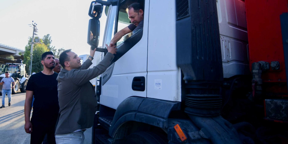 Gri ceketli bir adam, beyaz bir temizlik kamyonunun şoför koltuğunda oturan sürücüyle el sıkışıyor. Kamyonun yanında siyah tişörtlü bir başka adam daha duruyor ve ikiliyi izliyor. Arka planda ise başka bir iş makinesi ve güneşli bir dış mekan görülüyor.