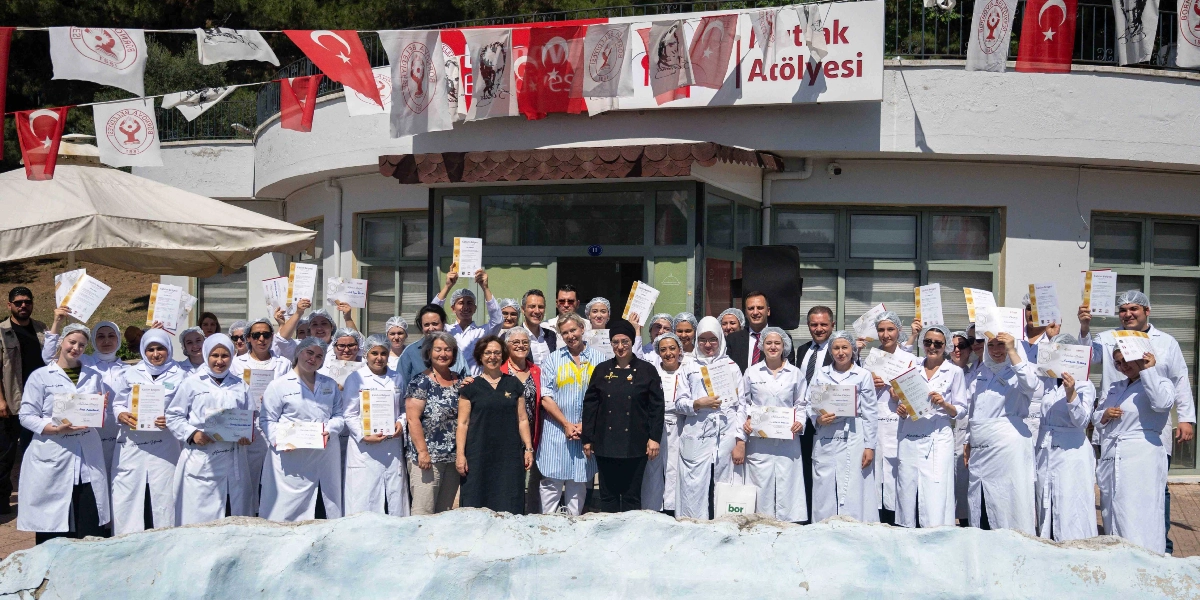 Bornova Belediyesi Mutfak Atölyesi önünde, beyaz önlükler giymiş ve ellerinde katılım belgelerini tutan kalabalık bir kursiyer grubu, eğitmenleri ve yetkililerle birlikte hatıra fotoğrafı çektiriyor. Binanın dış cephesi Türk bayrakları ve Bornova Belediyesi flamalarıyla süslenmiş durumda.