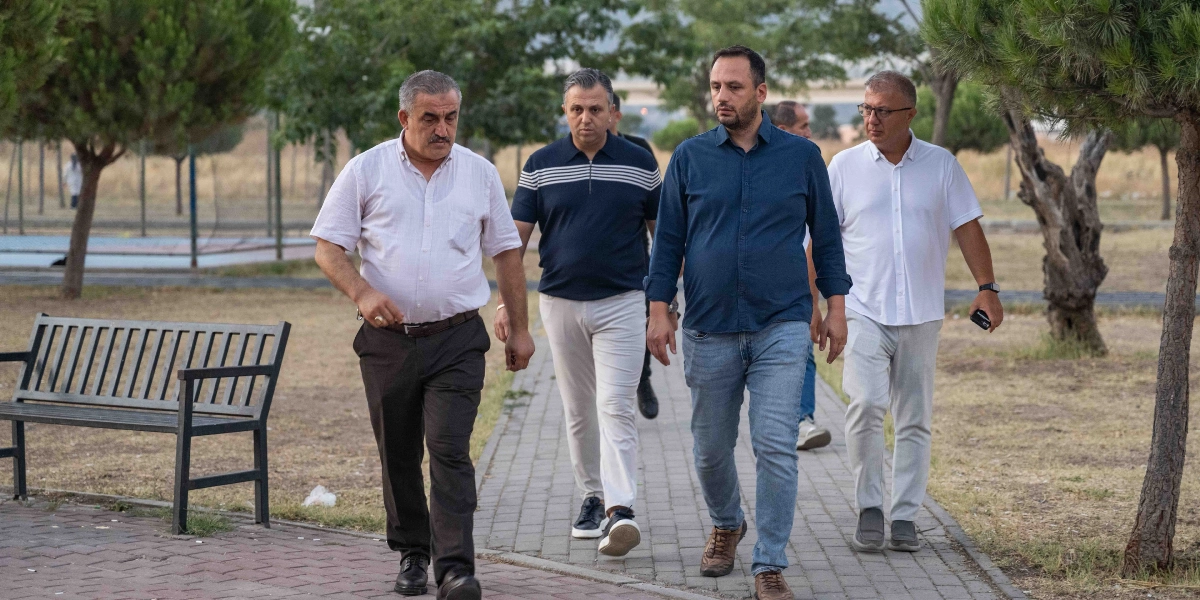 Açık havada, park içindeki taş döşeli bir yolda yan yana yürüyen dört erkek. En soldaki kişi beyaz kısa kollu gömlek ve koyu renk pantolon giyiyor. Yanındaki kişi lacivert çizgili polo yaka tişört ve açık renk pantolonla ilerliyor. Üçüncü kişi lacivert gömlek ve kot pantolon giymiş, en sağdaki kişi ise beyaz polo yaka tişört ve açık renk pantolonla onlara eşlik ediyor. Arka planda ağaçlar ve boş bir bank bulunuyor.