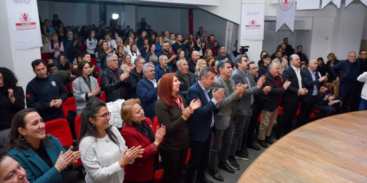 Konferans salonunda ayakta duran ve sahneye doğru dönerek alkışlayan kalabalık bir izleyici grubu. Salonun duvarlarında 