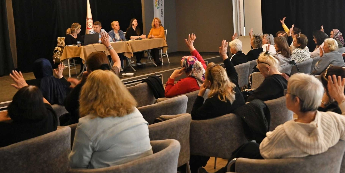 Kapalı bir salonda düzenlenen toplantıda, sahnedeki masada oturan beş kişilik bir panelist grubu ve onları dinleyen, sorular sormak için ellerini kaldıran çok sayıda kadın katılımcı yer alıyor. Katılımcılar gri koltuklarda oturuyor ve toplantı salonunun ön tarafında Bornova Kent Konseyi'ne ait olduğu anlaşılan bir flama ve bilgilendirme panosu bulunuyor.