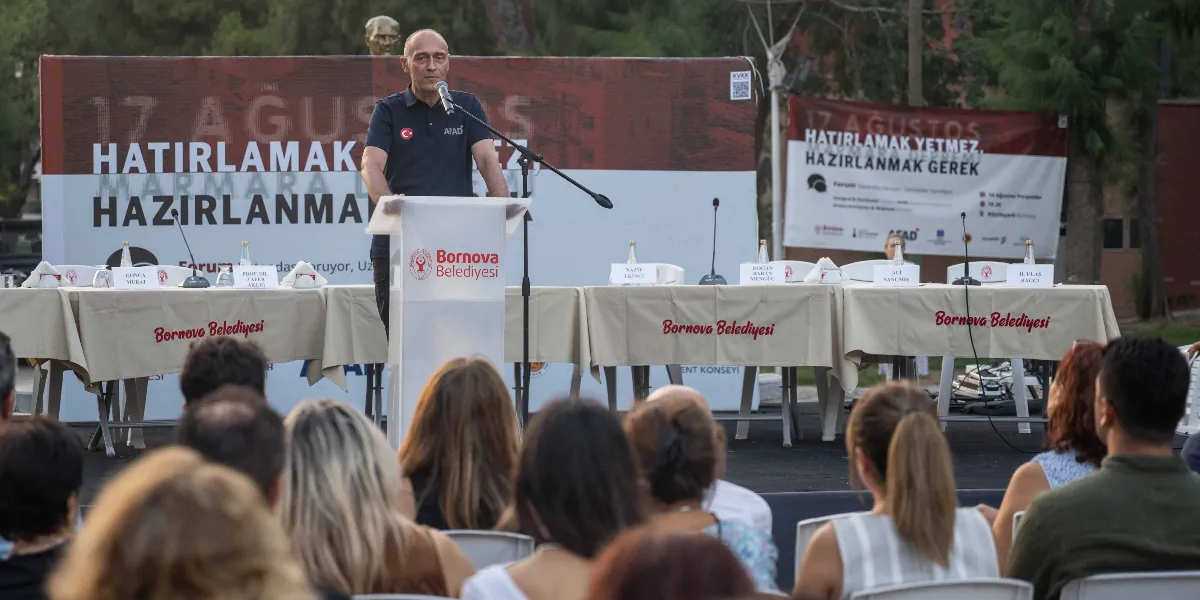 Bornova Belediyesi tarafından düzenlenen bir etkinlikte, üzerinde AFAD logosu bulunan lacivert polo yaka tişörtlü bir konuşmacı, kürsüde hitap ediyor. Konuşmacının arkasında 