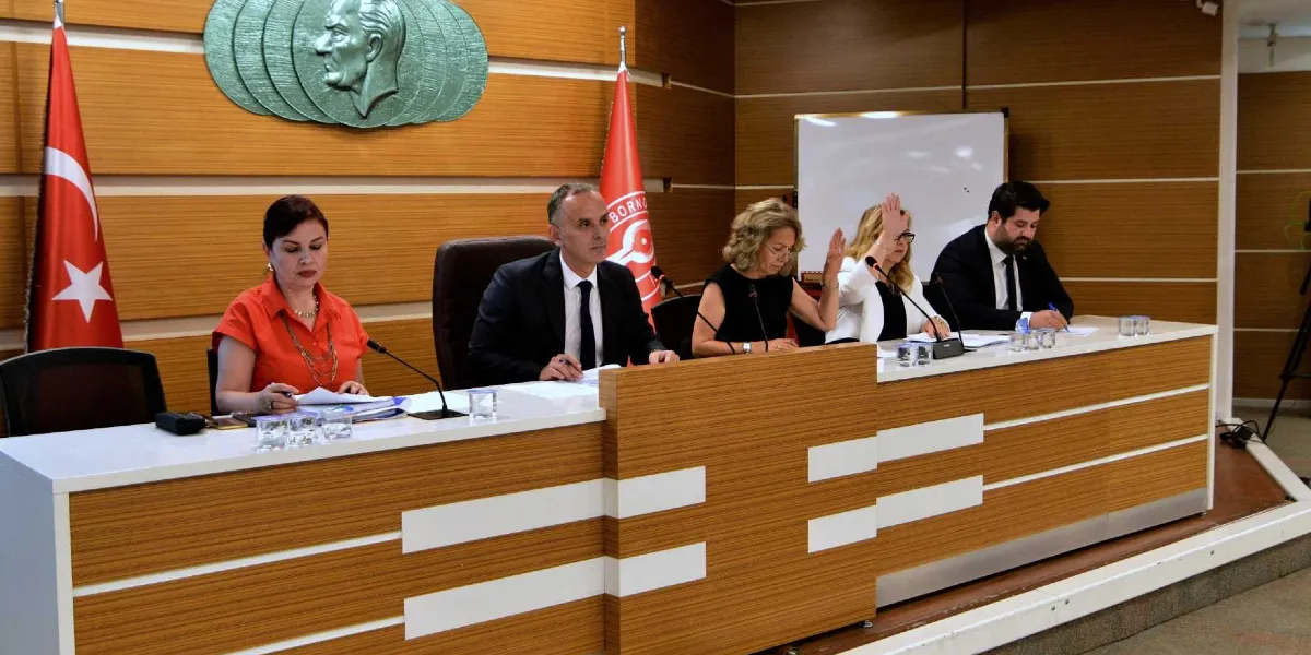 Ahşap kaplamalı bir toplantı masasında yan yana oturan dört kişi, resmi bir oturum gerçekleştiriyor. Masanın arkasındaki duvarda Atatürk kabartması ve Bornova Belediyesi'nin logosunun yer aldığı bir bayrak bulunuyor. Soldan sağa doğru turuncu bluzlu bir kadın, takım elbiseli bir erkek, siyah kıyafetli bir kadın ve beyaz ceketli bir kadın oturuyor; beyaz ceketli kadın elini kaldırarak oy kullanıyor veya söz istiyor. En sağda ise takım elbiseli bir erkek not alıyor. Masanın üzerinde mikrofonlar ve su bardakları yer alıyor.