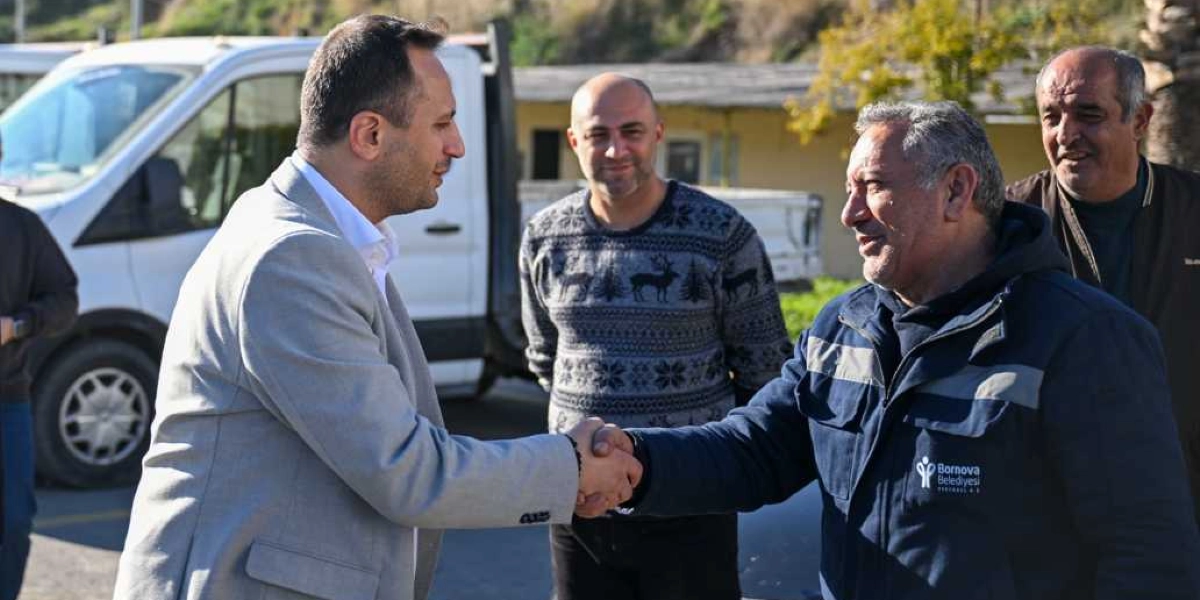 Açık renk ceket giymiş bir yetkili, üzerinde Bornova Belediyesi logosu bulunan lacivert bir iş kıyafeti giyen bir çalışanla tokalaşıyor. İkiliye arkalarında duran iki kişi daha eşlik ediyor ve arka planda beyaz bir kamyonet görülüyor.