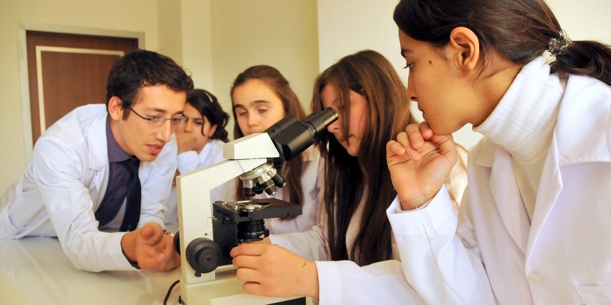 Beyaz laboratuvar önlüğü giymiş bir öğretmen ve üç öğrenci, bir laboratuvar ortamında mikroskop başında çalışıyor. Öğretmen, mikroskobu inceleyen öğrencilere rehberlik ederken, öğrenciler dikkatle mikroskoba odaklanmış durumda.