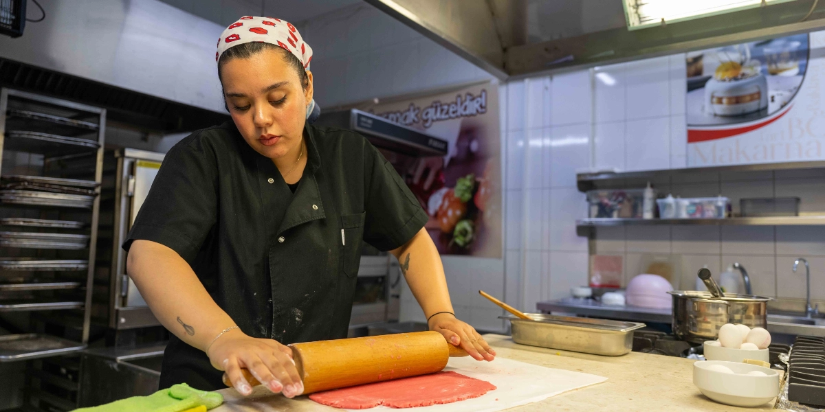 Profesyonel mutfak önlüğü giymiş, başında kırmızı dudak desenli bir bandana bulunan genç bir kadın, tezgah üzerinde merdane ile kırmızı renkli bir hamuru açıyor. Arka planda paslanmaz çelik mutfak ekipmanları, raflar ve üzerinde 