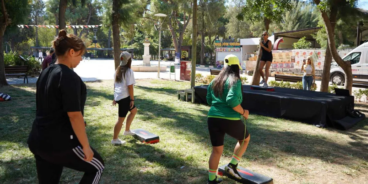 Ağaçlık bir park alanında, step tahtaları üzerinde egzersiz yapan üç kadın ve onları yönlendiren bir eğitmen yer alıyor. Siyah spor kıyafetli bir eğitmen, siyah örtüyle kaplı küçük bir platform üzerinde durarak hareketleri gösterirken, katılımcılar çimenlik alana yerleştirilmiş step tahtaları üzerinde ona eşlik ediyor. Arka planda bir dondurma standı, ağaçlar ve parkın genel görünümü bulunuyor.