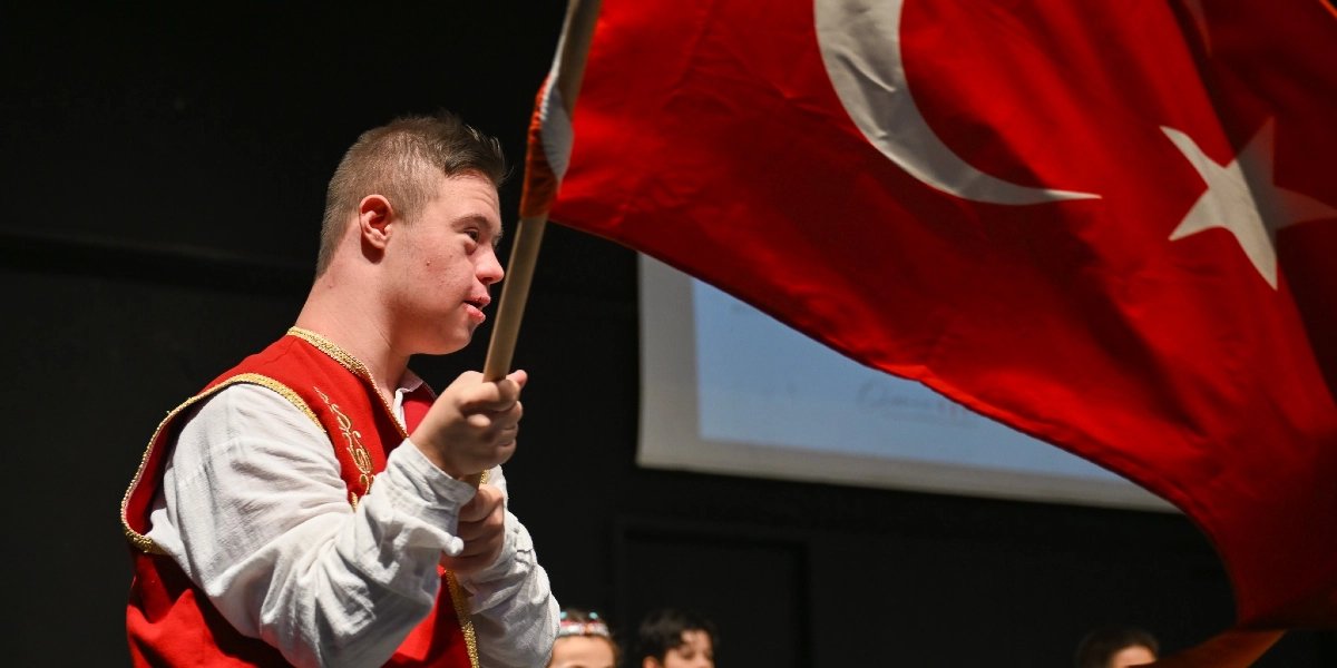 Geleneksel kırmızı yelek ve beyaz gömlek giymiş Down sendromlu genç bir erkek, elinde büyük bir Türk bayrağı tutarak sahne üzerinde gururla duruyor. Arka planda karanlık bir sahne düzeni ve kısmen görünen bir projeksiyon perdesi yer alıyor.
