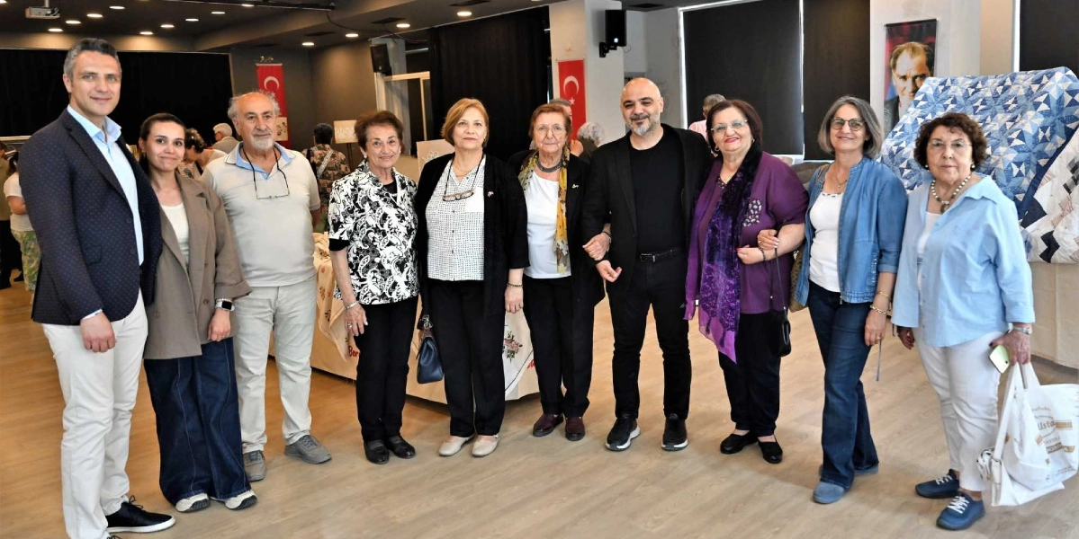 Atatürkçü Düşünce Derneği etkinliğinde yan yana duran, farklı yaş gruplarından dokuz kişilik bir grup. Gruptaki kadınlar ve erkekler günlük kıyafetler içinde, gülümseyerek kameraya bakıyor. Arka planda Türk bayrakları, Mustafa Kemal Atatürk'ün bir portresi ve sergilenen el işi ürünler ile patchwork bir örtü bulunuyor. İç mekan, aydınlık ve geniş bir salon görünümünde.