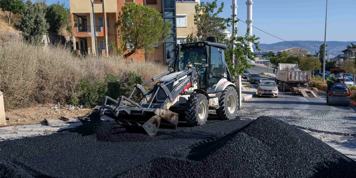 Bornova Belediyesi ekipleri, bir Hidromek marka iş makinesi kullanarak yol üzerinde asfalt serim ve düzenleme çalışması yapıyor. Ön planda büyük bir asfalt yığını bulunurken, arka planda yolun devamında çalışan bir silindir ve trafik akışının devam ettiği bir cadde görülüyor.